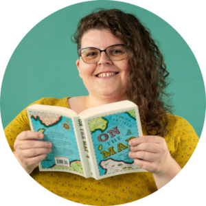 Claire Birnie is holding a map book and smiling at the camera. She is wearing a yellow jumper, has brown curly hair and glasses.