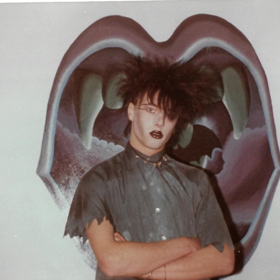 Paul is seen here with long dark hair across his eyes and dramatic makeup with dark lips, pale skin and dark eyes, He is wearing a grey shirt and stood in front of a  painted depicting a mouth.