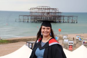 A young woman in a graduation gown is smiling at the camera with a background of a beach