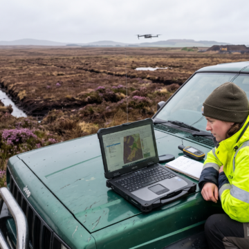 An ecologist in yellow high vis is looking at a laptop which is balanced on the bonnet of a car. They are in a heathland.