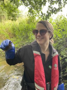 Carol is smiling and holding up a crayfish. She has dark hair, sunglasses and is wearing a red buoyancy aid.