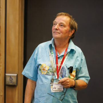 Dr M is wearing a blue shirt and a red lanyard. He has brunette hair and is looking to the side with a calm, happy expression.